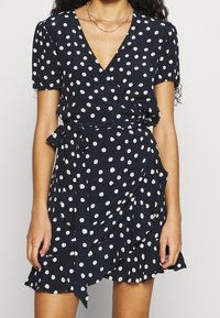 Námornický zavinovací šaty s bílými puntíky, krátkými rukávy, výstřihem do V, volánkovým lemem a detailem vázání v pase. Hladká textura látky.