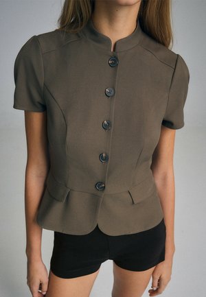 Woman wearing a fitted olive green short-sleeve jacket with black buttons and black shorts against a plain background.