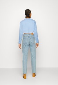 Jasnoniebieska krótka koszula, z długimi rękawami i kołnierzykiem, w zestawie z jasnoniebieskimi dżinsami o wysokim stanie. Modelka nosi brązowe buty na obcasie.