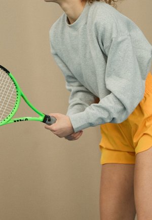 Person wearing gray sweatshirt and yellow shorts holding a green tennis racket in a ready position against a plain background.
