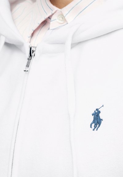 Felpa con cappuccio bianca con cerniera indossata sopra una camicia con colletto a righe blu, con un piccolo logo ricamato di un giocatore di polo blu sul petto.