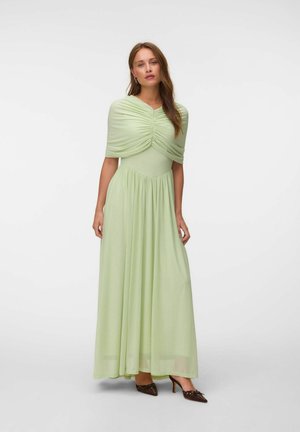 Femme portant une longue robe vert clair froncée et des talons pointus marron, debout sur un fond blanc uni.