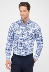 Man in een blauwe en witte overhemd met lange mouwen met ingewikkelde pastorale scènes, gecombineerd met donkerblauwe broek, staande met één hand in zijn zak.