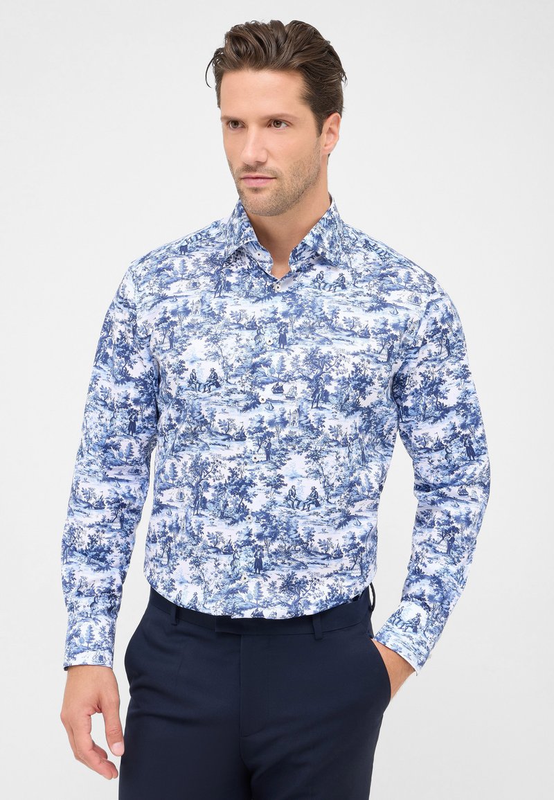 Man in een blauwe en witte overhemd met lange mouwen met ingewikkelde pastorale scènes, gecombineerd met donkerblauwe broek, staande met één hand in zijn zak.