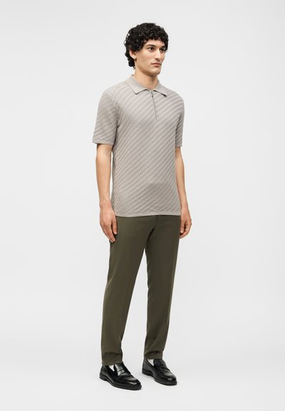 Άνδρας με σγουρά μαλλιά που φοράει μια μπεζ κοντομάνικη πόλο μπλούζα με υφή, ελιά παντελόνια και μαύρα επίσημα παπούτσια, στέκεται σε απλό φόντο.