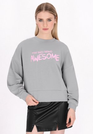 Kobieta o prostych blond włosach, ubrana w szarą bluzę z różowym napisem „Too busy being awesome” oraz czarną skórzaną mini spódniczkę.