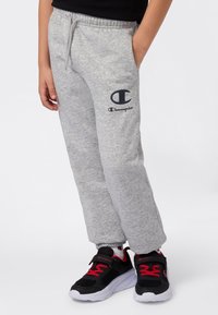 Pantalones de chándal grises claros con una cintura elástica y bolsillos, con un logotipo de Champion en azul marino. Combinados con zapatillas negras y acentos rojos.