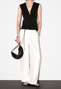 Svart ärmlös topp med djup V-ringning och knappar fram, kombinerad med löst sittande vita byxor, håller en svart halvmåneformad handväska. Silverarmband.