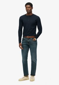 Marineblaues Langarm-Henley-Shirt mit Knöpfen, kombiniert mit blauen Jeans, einem braunen Leder-Gürtel und hellen Sneakers.