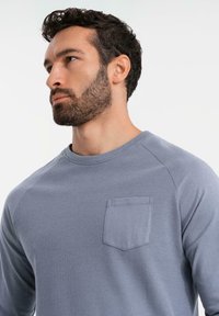 Vaaleansininen pitkähihainen paita, jossa on teksturoitu vartalo ja sileät raglan-hihat, sekä pieni tasku vasemmalla puolella.