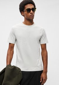 Svetlo šedé tričko s krátkym rukávom, okrúhly výstrih, s jemným logom. Model má tmavé slnečné okuliare a drží olivovo zelenú bundu.