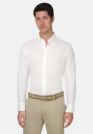 Jeune homme portant une chemise blanche boutonnée rentrée dans un pantalon beige avec une ceinture tressée beige, debout devant un fond uni.