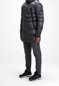 Man in een lange zwarte pufferjack met capuchon, grijze broek en zwarte New Balance sneakers, staand op een witte achtergrond.