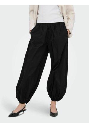 Kvinde iført løse sorte bukser med elastisk manchet, hvid top, beige blazer og sorte lavhælede slingback-sko, stående med hænderne i lommerne.