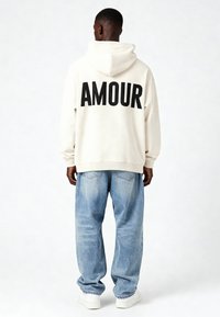 Kreminė hoodie su užrašu "AMOUR" juoda spalva ant nugaros, derinama su šviesiai mėlynomis plačiomis kelnėmis ir baltais sportbačiais. Laisvas kirpimas ir minkšta tekstūra.