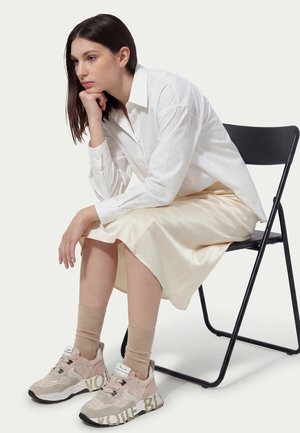 Femme aux cheveux foncés, vêtue d'une chemise blanche, d'une jupe beige, de chaussettes beiges et de baskets roses, assise sur une chaise pliante noire, le menton reposant sur son poing.