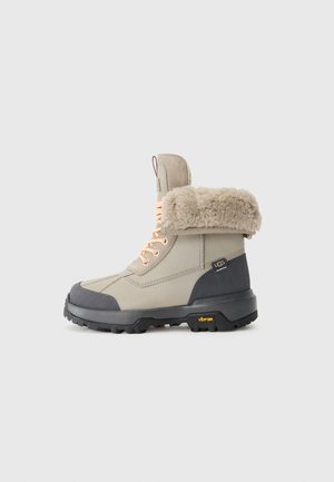 Beige vinterstøvel med grå detaljer, kunstpels krage og oransje snøring. Har gummisåle med Vibram-logo for bedre grep.