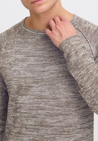 Pull en tricot à manches longues et texture, de couleur marron avec un motif chiné. Il présente un col et des poignets côtelés, avec une coupe décontractée.