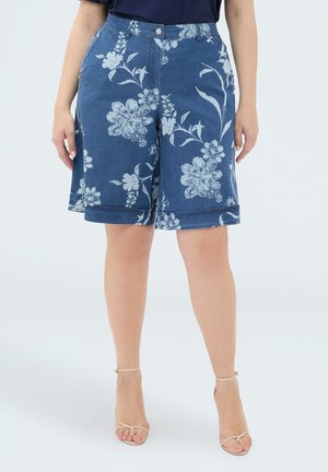 Niebieskie krótkie spodenki do kolan z białym kwiatowym nadrukiem, noszone z beżowymi sandałami na paski i ciemną bluzką, pokazane na stojącej osobie.