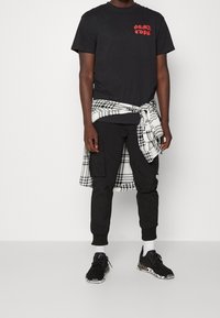 Černé tričko s červeným textem, spárované s černými cargo kalhotami. Bílo-černá kostkovaná košile uvázaná kolem pasu. Černé tenisky s vzorovanou podešví.