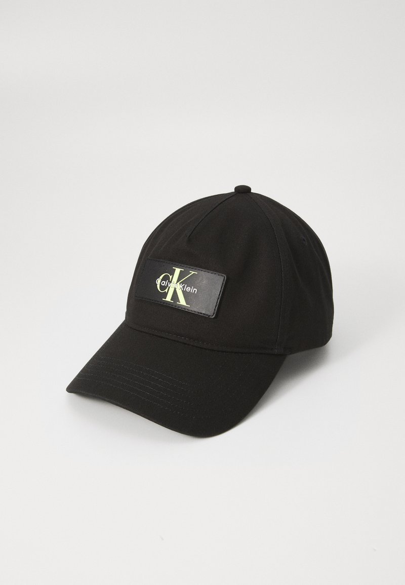 Zwarte honkbalpet van stof, voorzien van een rechthoekig logo-opgenaaid met groene tekst. Het ontwerp omvat een gestikte rand en ventilatiegaten.