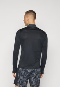 Maglietta sportiva nera a maniche lunghe con colletto alto, texture liscia e design aderente, abbinata a pantaloni corti scuri decorati con grafiche simili a pixel.