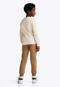 Maglione crema con orlo e polsini a coste, abbinato a pantaloni della tuta beige. Sneakers bianche con dettagli blu navy e rossi. Bambino che guarda lontano dalla macchina fotografica.