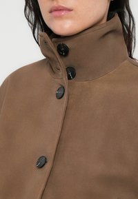 Καφέ σουέντ μπουφάν με ψηλό γιακά, με μαύρα κουμπιά και απαλή, λεία υφή. Κοντινή λήψη του επάνω μέρους του ρούχου.