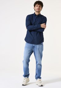 Marineblaues Button-Up-Hemd mit strukturiertem Stoff, langen Ärmeln, kombiniert mit hellblauen Jeans und beigen Sportschuhen. Das Modell steht mit verschränkten Armen.