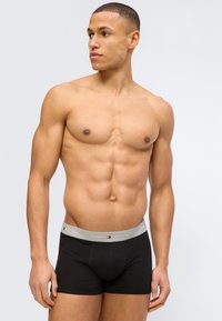 Αθλητικός νεαρός άνδρας που φοράει μαύρα ανδρικά εσώρουχα Tommy Hilfiger με γκρι μέση, κοιτάζοντας προς τα πλάγια με φόντο ένα απλό ανοιχτόχρωμο υπόβαθρο.