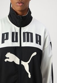 Mustvalge lukuga jakk, millel on suur must "PUMA" logo ja ees valge hüppev puma graafika. Sile, sportlik kangas.