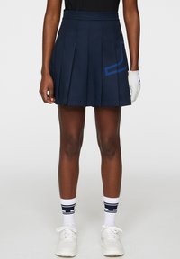 Marinblå plisserad golfkjol med en kontrasterande blå grafisk design. Kombinerad med vita sportskor och randiga strumpor, med en handske på ena handen.