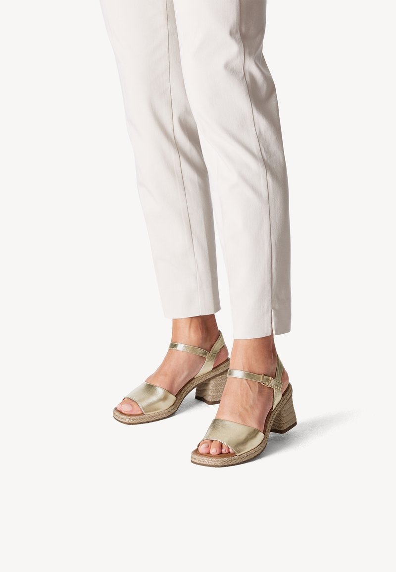 Gold metallic sandals with ankle strap and open toe, featuring a textured woven wedge heel. Paired with tailored white pants.