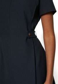 Marinblå klänning med korta ärmar, en åtsittande siluett och en sidodetalj med en knapp. Tillverkad av ett mjukt tyg med en subtil textur.