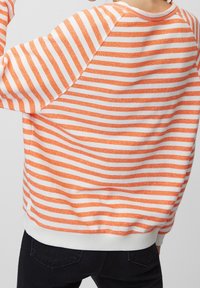 Oranssi-valkoraidallinen collegepaita rentolla istuvuudella, resorilla hihansuissa ja helmassa sekä pehmeällä tekstuurilla, jossa on raglan-hiha.