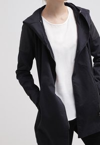 Chaqueta con capucha de color marino, que presenta un cierre de cremallera frontal, dos bolsillos laterales, hombros acanalados y un corte relajado, llevada sobre una camiseta blanca.