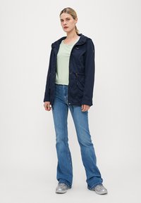 Veste bleu marine avec capuche, taille cintrée et poches latérales, portée sur une chemise vert clair et un jean évasé bleu. Baskets argentées exposées.