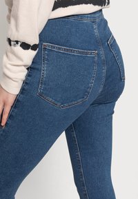 Jeans skinny de cintura alta, feitos de denim azul, com dois bolsos traseiros e detalhes de costura discretos. Textura suave com um formato ajustado.
