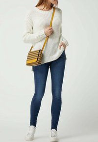 Mujer vestida con un suéter de punto blanco, jeans ajustados azules, zapatillas blancas y un bolso cruzado marrón a cuadros con strap amarillo.