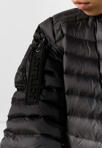 Giacca nera imbottita con trapuntatura orizzontale, dotata di una zip laterale e di un patch logo testurizzato sulla manica. Materiale liscio e riflettente.