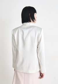 White blazer with a structured silhouette. Smooth texture, long sleeves, and a subtle sheen. Back features a centered seam.