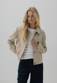 Jasno beżowa, lekka kurtka z kołnierzem, dwoma przednimi kieszeniami i zapięciem na guziki, zestawiona z białą koszulą i ciemnoniebieskimi dżinsami.