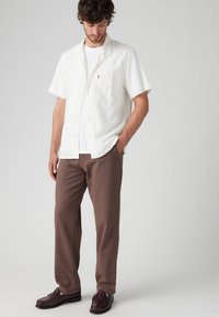 Witte korte mouwen textuur shirt over een witte t-shirt, gecombineerd met bruine broeken en donkere instappers, wat een gelaagde casual look creëert.