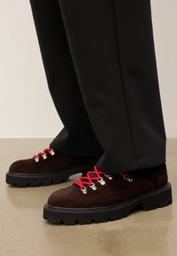 Pantofi din daim maro cu talpă din cauciuc negru gros, șireturi roșii și ochiuri din metal. Asortați cu pantaloni negri.