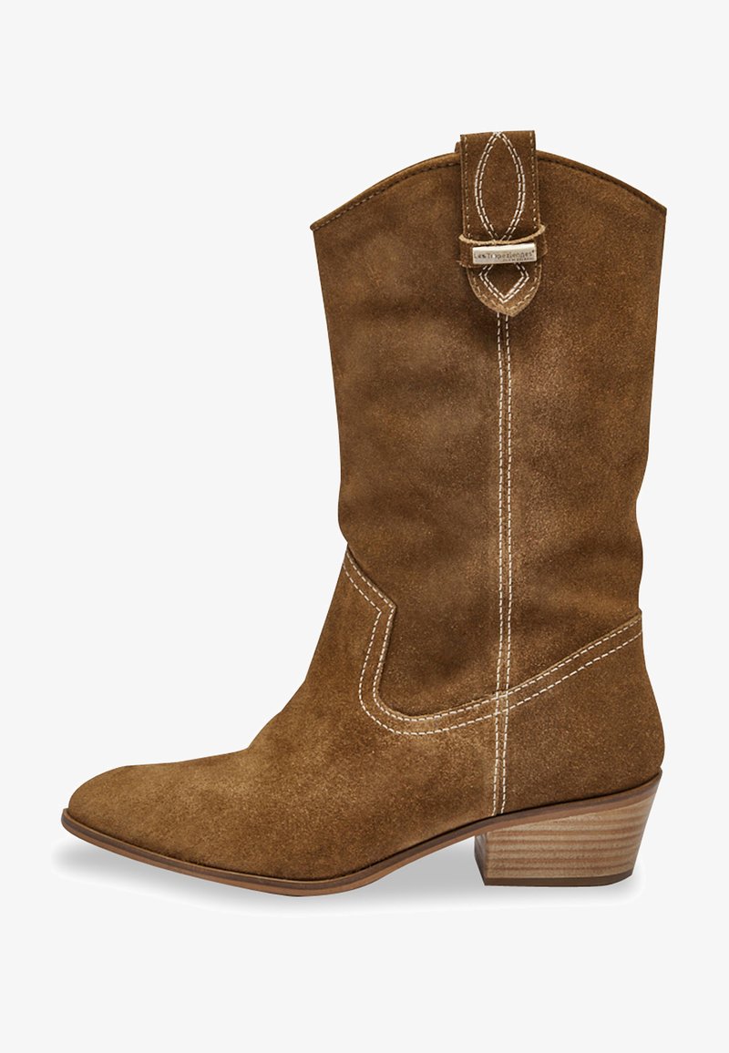 Botte western en daim marron avec un bout effilé, des coutures courbées sur les côtés et un petit talon empilé. Elle présente une sangle décorative en haut.