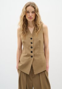 Gilet senza maniche color sabbia con scollo a V profondo e bottoni neri sul davanti. Due tasche laterali. Tessuto liscio con vestibilità su misura.