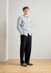 Camisa de botones a rayas en azul y blanco, combinada con pantalones holgados negros y zapatos grises, de pie sobre un suelo de madera.