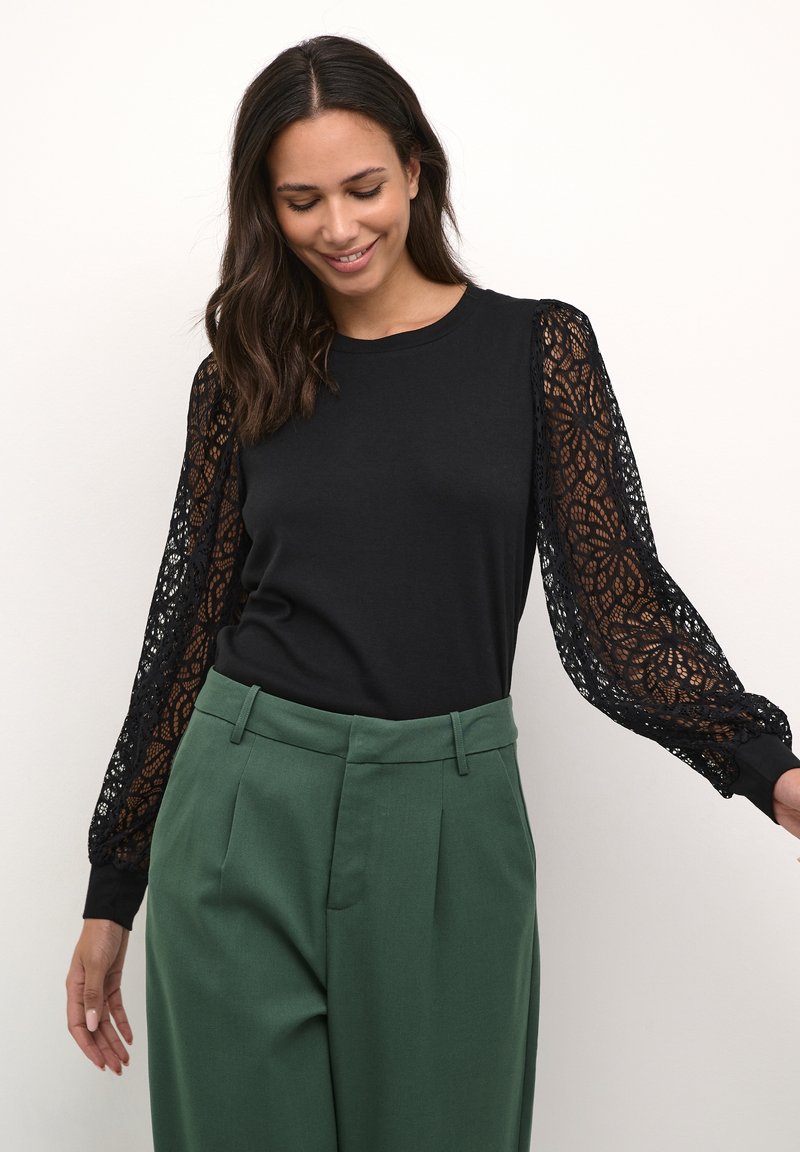 Femme portant un haut noir à manches longues en dentelle et un pantalon vert taille haute, souriant les yeux fermés sur un fond uni.