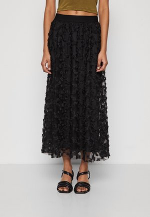 Maxi falda de encaje negro con flores, con una cinturilla elástica, que presenta una textura en capas y una superposición transparente. Combinada con sandalias negras de tiras.