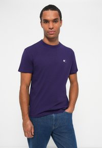 T-shirt di cotone viola con colletto rotondo e maniche corte. Presenta un piccolo logo stampato in bianco sul lato sinistro del petto. Indossato con jeans blu.